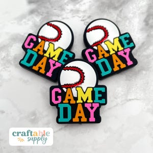 May include: Three baseball-themed charms with the words "GAME DAY" in colorful letters. Each charm features a white baseball with red stitching and a black border. The charms are designed for crafting and are ideal for sports fans.