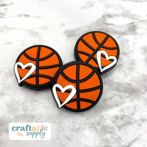 May include: Three orange and black basketball charms with white hearts. The charms are made of silicone and are perfect for adding a sporty touch to your crafts.