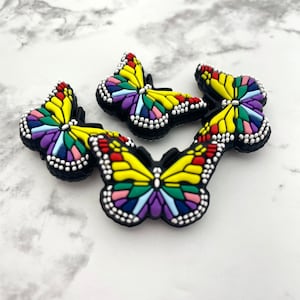 May include: Three colorful butterfly shoe charms with a yellow, red, blue, green, and purple mosaic design. The charms are made of rubber and have a black background.