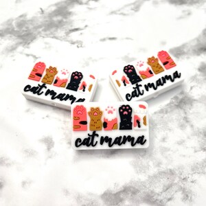 May include: Three white rectangular silicone charms with the text "cat mama" and a row of colorful cat paw prints.