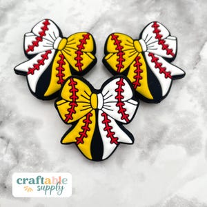 May include: Three baseball-themed bow charms. Each charm features a yellow center, white and black accents, and red stitching, resembling a baseball. The charms are arranged on a light gray surface. The text "craftable supply" is visible.