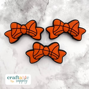 May include: Three orange bow-shaped charms with black outlines, resembling basketballs. The charms are arranged on a light gray and white speckled surface. The charms are likely for crafting or jewelry making.