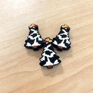 May include: Three black and white cow print Christmas tree ornaments with brown cowboy hats. The ornaments are made of a soft, pliable material.