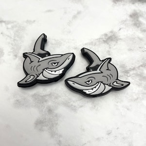 Puede incluir: Dos dijes grises con forma de tiburón con detalles en blanco y negro. Los tiburones tienen una expresión sonriente y están diseñados con una aleta y branquias. Estos dijes son probablemente para zapatos o accesorios.