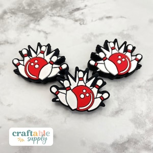 May include: Three bowling-themed charms. Each charm features a red bowling ball with white pins and a black outline. The charms are arranged on a light gray and white marbled surface. The text "craftable supply" is in the lower left corner.