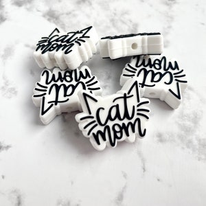 May include: Four white silicone cat-shaped charms with black text. The charms read "Cat Mom" and "Meow Cat".