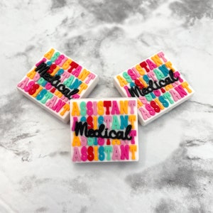 May include: Three square charms with the words "Medical Assistant" in colorful block letters. The text is in a repeating pattern of orange, pink, green, and blue on a white background. The words "Medical Assistant" are in black cursive.