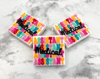 Op de afbeelding: Drie vierkante bedels met de woorden "Medical Assistant" in kleurrijke blokletters. De tekst staat in een herhalend patroon van oranje, roze, groen en blauw op een witte achtergrond. De woorden "Medical Assistant" zijn in zwart cursief.