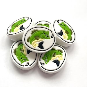 May include: A collection of white, oval-shaped charms featuring a green fish design with black and yellow accents. The fish is set against a white background within a black-lined oval. These charms are likely for shoes or accessories.