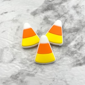 May include: Three white, yellow, and orange candy corn charms. The charms are shaped like a traditional candy corn.