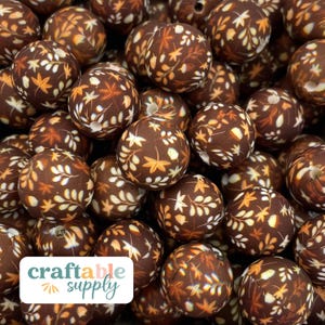 May include: A close-up of a pile of brown silicone beads with a white and orange leaf pattern. The beads are approximately 2 cm in diameter. The beads are arranged in a random pattern. The beads are for crafting and are sold by the company "craftable supply".