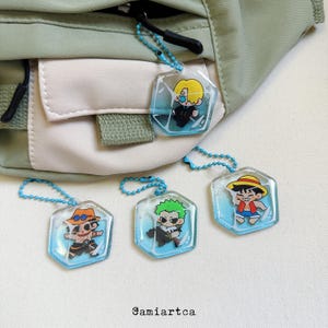 May include: Four hexagonal acrylic keychains with anime character designs. Each keychain has a blue chain. The keychains are attached to a green and beige bag. The image includes the text "@amiartca".