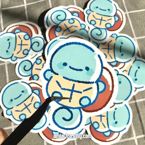 Pode incluir: Uma cole&ccedil;&atilde;o de autocolantes de desenhos animados com uma personagem azul clara semelhante a uma tartaruga com uma carapa&ccedil;a amarela e um rosto sorridente. Os autocolantes t&ecirc;m uma borda branca e um design de carapa&ccedil;a vermelho e castanho. O texto "amiartea.etsy.com" &eacute; vis&iacute;vel.