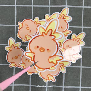 Pode incluir: Uma cole&ccedil;&atilde;o de adesivos de p&aacute;ssaros de desenho animado laranja e amarelo. Os adesivos apresentam um design de p&aacute;ssaro fofo com um corpo redondo, asas e uma crista amarela. Os adesivos est&atilde;o em um fundo de grade cinza. O nome do artista, "Camiartea", &eacute; vis&iacute;vel.