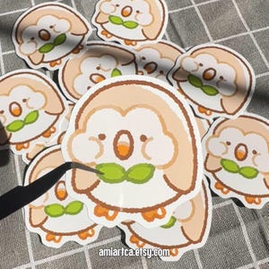 Pode incluir: Uma cole&ccedil;&atilde;o de autocolantes de desenhos animados com uma criatura redonda e fofa com um corpo castanho claro, bico laranja e folha verde. Os autocolantes est&atilde;o a ser manuseados com pin&ccedil;as. O URL da loja Etsy "amiartca.etsy.com" &eacute; vis&iacute;vel.