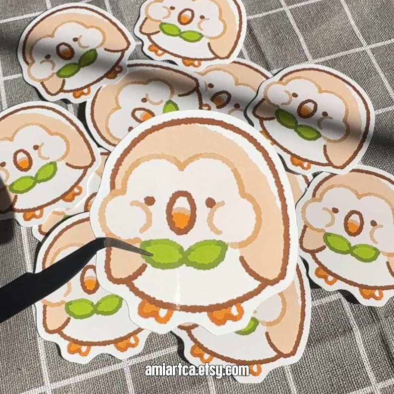 Pode incluir: Uma cole&ccedil;&atilde;o de autocolantes de desenhos animados com uma criatura redonda e fofa com um corpo castanho claro, bico laranja e folha verde. Os autocolantes est&atilde;o a ser manuseados com pin&ccedil;as. O URL da loja Etsy "amiartca.etsy.com" &eacute; vis&iacute;vel.