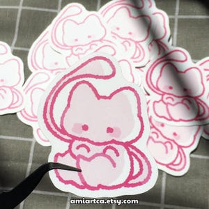 Pode incluir: Uma cole&ccedil;&atilde;o de autocolantes cor-de-rosa e brancos com um design de gato de desenho animado. O gato tem um corpo arredondado, uma cauda longa e bochechas cor-de-rosa. Os autocolantes est&atilde;o numa superf&iacute;cie cinzenta xadrez. O texto "amiartca.etsy.com" &eacute; vis&iacute;vel.