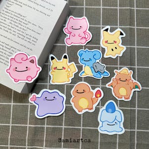 Könnte beinhalten: Eine Sammlung bunter Cartoon-Aufkleber mit verschiedenen Charakteren. Die Aufkleber zeigen ein rosa katzenartiges Wesen, ein gelbes Pikachu, ein blaues Lapras und andere. Die Aufkleber befinden sich auf einer karierten Oberfläche, mit einem teilweise sichtbaren Buch.