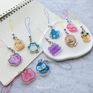 May include: A collection of ten colourful acrylic charms featuring various cartoon characters, each attached to a small metallic jump ring and a thin nylon cord loop in matching colours. The charms display diverse shapes and vibrant illustrations.