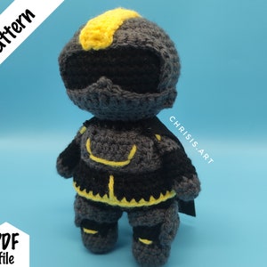 May include: Crocheted amigurumi toy of a black and yellow superhero with a cape. The toy is wearing a helmet and has a detailed design.