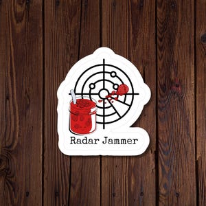 May include: A white sticker with a black radar graphic and a jar of red jam with a spoon. The text "Radar Jammer" is below the jar and radar graphic. The sticker is on a wooden background.