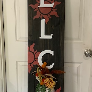 May include: A wooden welcome sign with the word "WELCOME" painted in white letters. Each letter is surrounded by a red sun with orange rays. A small potted plant with green leaves and orange flowers is hanging from the sign.