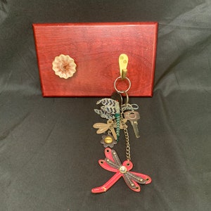 May include: A wooden key holder with a brass hook and a decorative ceramic knob. A key chain with a dragonfly charm and several keys hangs from the hook.