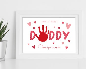 Può includere: Stampa incorniciata con bordo bianco. La stampa presenta la parola "DADDY" in lettere rosse, con un'impronta della mano rossa che sostituisce la lettera "A". Sono inclusi anche i testi "Happy Valentine's" e "I love you so much", insieme a cuori rosa.