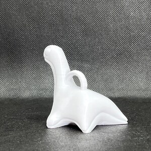 May include: White ceramic dinosaur figurine with a loop on the back for hanging. The dinosaur is in a sitting position with its tail curled around its legs.