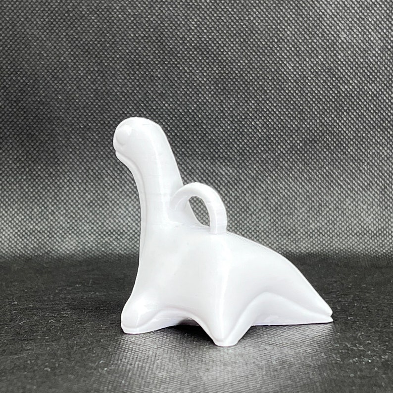 May include: White ceramic dinosaur figurine with a loop on the back for hanging. The dinosaur is in a sitting position with its tail curled around its legs.