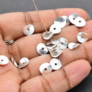 May include: Silver metal disc beads with a textured finish and a central hole. Some beads are strung on a copper wire. The beads are held in a hand, indicating their use in jewellery making or crafts.
