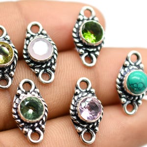 Pode incluir: Seis pingentes de prata com pedras preciosas de diferentes cores. Cada pingente tem dois laços para prender a uma corrente ou pulseira. As pedras preciosas são verdes, rosa, brancas, amarelas e turquesa.