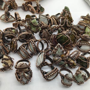 May include: A collection of rings featuring various natural stones wrapped in copper wire. Each ring displays a unique, irregularly shaped gemstone secured by intricate wirework coils and bands, showcasing earthy tones of green, gray, and brown.