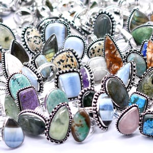 May include: A collection of gemstone rings with various shapes and colors, including turquoise, purple, and green stones. The rings have silver-colored settings with a beaded border, creating a vintage or bohemian aesthetic.