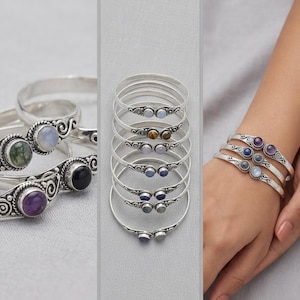Può includere: Una collezione di bracciali rigidi in metallo color argento con intricati motivi a volute e due pietre preziose rotonde a cabochon incastonate alle estremità. I bracciali sono mostrati in varie disposizioni, con pietre di diversi colori tra cui viola, blu e verde.