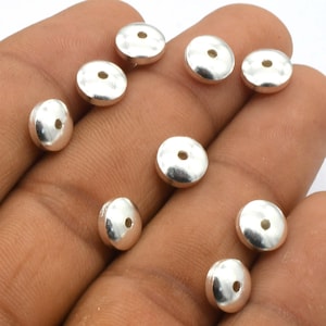 Puede incluir: Una colección de cuentas de metal plateadas con un agujero en el centro. Las cuentas son redondas y planas, y están dispuestas en un patrón aleatorio.