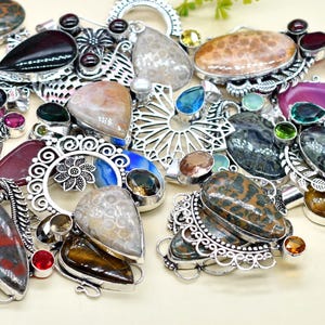 May include: A collection of gemstone pendants with silver-colored metal settings. The pendants feature various shapes and colors, including oval, teardrop, and round stones in shades of green, purple, brown, and blue. Some have intricate filigree designs.