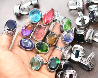 May include: A collection of gemstone rings in various colors, including blue, red, green, and purple. The rings have ornate silver bands and feature oval, rectangular, and round-cut stones. The rings are displayed on a hand.