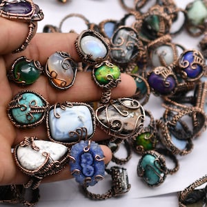 May include: A selection of handcrafted rings, each featuring a unique gemstone set within copper wire. The rings showcase a variety of stones, including turquoise, amethyst, and moonstone. The gemstones are in shades of blue, green, white, and purple.