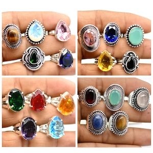 Op de afbeelding: Een verzameling van 12 zilveren ringen met verschillende edelsteen bezettingen. De ringen tonen een verscheidenheid aan kleuren, waaronder groen, rood, blauw, geel, roze en paars. De edelstenen zijn gezet in verschillende stijlen, waaronder peervormig, ovaal en rond.