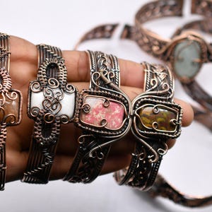 May include: A collection of handcrafted copper and silver bracelets. The bracelets feature intricate wirework designs and are adorned with various gemstones, including a light brown, white, pink, and green stone. The bracelets are displayed on a white background.