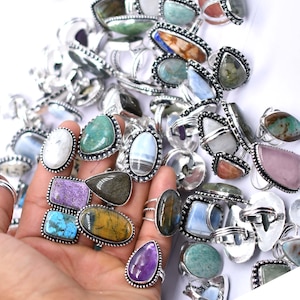 Puede incluir: Una colección de anillos de piedras preciosas con varias formas y colores, incluyendo piedras turquesas, moradas y marrones. Los anillos tienen engastes de metal plateado y se muestran sobre un fondo blanco.