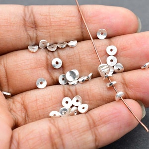 May include: A close-up of a hand holding a collection of silver-toned metal beads. The beads are round and flat, with a hole in the centre. The beads are arranged in a loose pile, with some of them strung on a thin wire.
