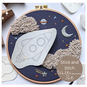Könnte beinhalten: Gesticktes Stickrahmen-Kunstwerk mit Weltraum-Thema. Der marineblaue Stoffhintergrund ist mit einer Rakete, Wolken, Sternen, einem Planeten und einem Mond verziert. Der Text "Stick and Stich Weltraum Set" ist ebenfalls enthalten.