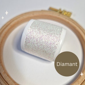 Könnte beinhalten: Eine Spule irisierendes Diamant-Garn mit weißer Kunststoffbasis liegt in einem hölzernen Stickrahmen. Das Garn hat ein schimmerndes, mehrfarbiges Aussehen. Das Wort "Diamant" befindet sich in einem braunen Kreis.