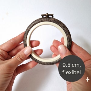 Könnte beinhalten: Ein flexibler Stickrahmen mit einem Durchmesser von 9,5 cm, mit einem äußeren Holzring und einem inneren weißen Ring. Der Rahmen wird in zwei Händen gehalten.