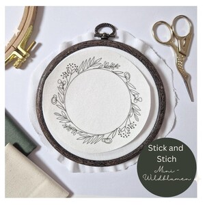 Könnte beinhalten: Ein Holz-Stickrahmen mit weißem Stoff im Inneren. Der Stoff hat eine schwarze Linienzeichnung eines Blumenkranzes. Der Text "Stick and Stich Mini - Wildblumen" ist unter dem Rahmen gedruckt.