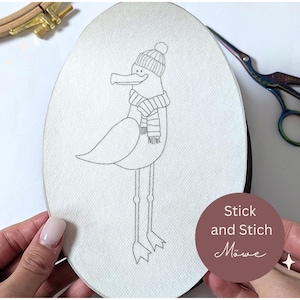 Könnte beinhalten: Gesticktes Design einer Ente mit gestreiftem Schal und Mütze. Die Ente steht auf langen Beinen. Der Text "Stick and Stich Möwe" befindet sich in einem braunen Kreis.