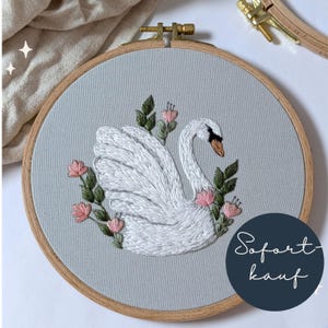 Könnte beinhalten: Gesticktes Schwan-Hoop-Art. Das Design zeigt einen weißen Schwan mit schwarzem Schnabel und Auge, umgeben von rosa Blüten und grünen Blättern. Das Kunstwerk ist auf einem hellgrauen Stoff angebracht und in einem Holzreifen gerahmt.