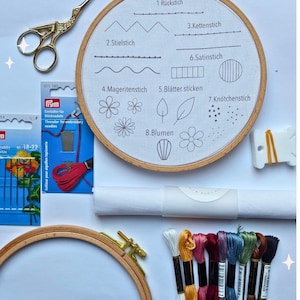 May include: Embroidery kit with a wooden hoop, scissors, needles, and colorful threads. A white fabric hoop displays stitch examples with German labels. Includes a needle threader and a roll of white fabric.